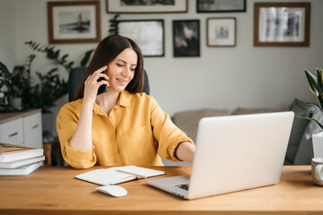 Smiling business woman talking on a mobile phone and using a laptop while working at home, looking at the screen, reading a message, a young female employee advises a client, telephone conversations