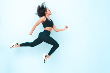 Fitness smiling black woman in sports clothing with afro curls hairstyle.She wearing sportswear. Young beautiful model with perfect tanned body.Female jumping and running in studio near blue wall