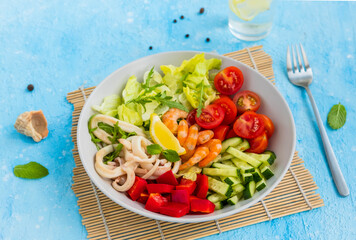 Healthy salad with fresh vegetables, shrimps and squid in a gray bowl on a light blue concrete background. Mediterranean cuisine.