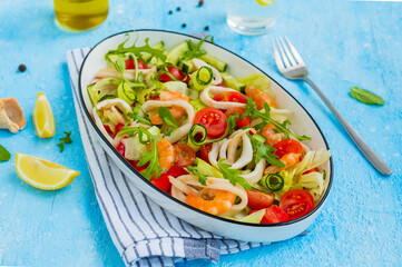 Healthy salad with fresh vegetables, shrimps and squid in a white oval bowl on a light blue concrete background. Mediterranean food.