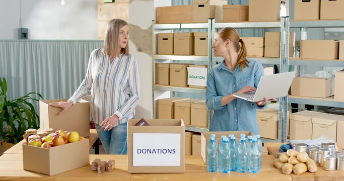 Caucasian young beautiful woman charity center owner speaking with senior female worker in warehouse and typing on laptop. Woman preparing and packing donation box, social help, charity concept