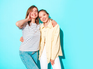 Two young beautiful blond smiling hipster female in trendy summer clothes. Sexy carefree women posing near blue wall in studio. Trendy and positive models having fun