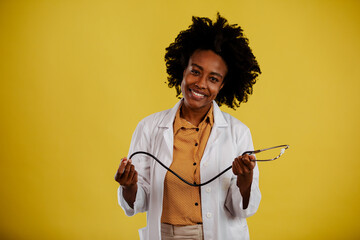 African American female doctor holding stethoscope and smiling while posing on yellow background.