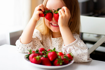  Cute little girl eating fresh strawberry in the kitchen. Healthy vitamin snack for kids. Ripe fresh berries. Harvest season. Natural vitamins .