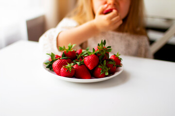  Cute little girl eating fresh strawberry in the kitchen. Healthy vitamin snack for kids. Ripe fresh berries. Harvest season. Natural vitamins .