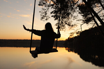 silhouette of a romantic young woman on a swing over lake at sunset. Young girl traveler sitting on the swing in beautiful nature, view on the lake