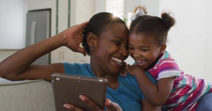 Happy african american mother and daughter sitting on sofa using digital tablet and laughing