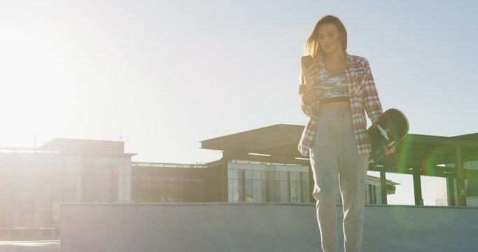 Caucasian woman, walking, using smartphone and holding skateboard at a skatepark