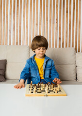 little boy chess player with brown hair and denim clothes is sitting on the sofa and playing chess in the room