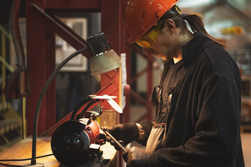 A mechanic wearing black gloves, yellow glasses and a hard hat processes a metal part on a grinding...