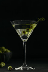 Glass of classic dry martini cocktail with olives on dark stone table against a black background.