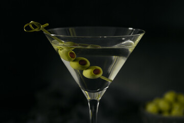Glass of classic dry martini cocktail with olives on dark stone table against a black background.