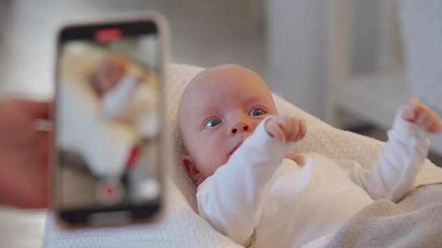 Mother filming her baby on a mobile phone camera at home, infant lying on white baby bouncer covered with gray blanket. High quality 4k footage