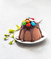 Chocolate curd dessert Easter, poured with icing and decorated with colorful candies on a plate on a linen napkin on a light gray background. Bright Holiday