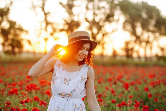 Portrait of a girl in a poppy field against a sunset background. Sunny photo of a woman in a straw hat and white dress. Brunette in flowers.