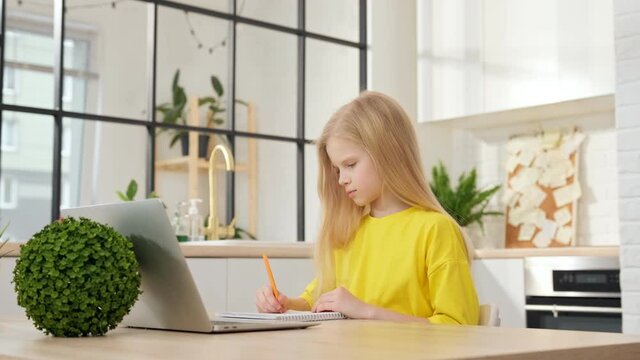 Blonde schoolgirl learns online using a laptop, taking notes in a notebook. Teenage schoolgirl girl is watching an Internet video course while sitting at her home table. Concept of distance learning.