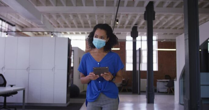 Mixed race businesswoman walking through corridor wearing facemask holding tablet