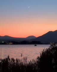 A crescent moon rising over Lake Kawaguchi and a blue and orange sky at dusk, with low mountains and a boat visible
