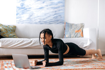 Beautiful young african american woman in sportswear, doing a plank on the floor at home, using a laptop, watching a video of fitness or yoga lessons, leading a healthy lifestyle, smiling