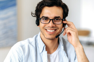 Close-up portrait of happy successful young hispanic man wearing glasses and a headset, freelance worker, call center or support service operator, looking at camera, smiling friendly