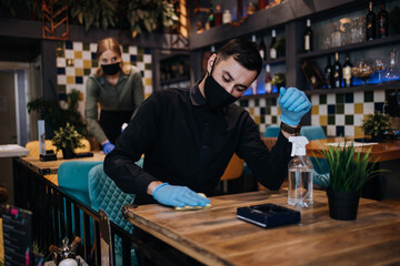 Young restaurant workers waiters cleaning and disinfecting tables and surfaces against Coronavirus pandemic disease. They are wearing protective face masks and gloves.
