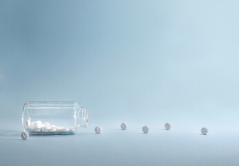 Transparent glass jar with white tablets rolling out of it. On a blue background.