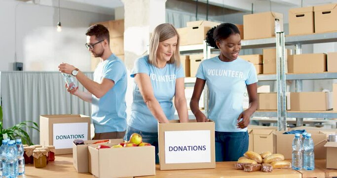 Mixed-race joyful male and female food bank volunteers putting food products in donation box working together as charity workers. People work in warehouse, social help for poor, humanitarian concept