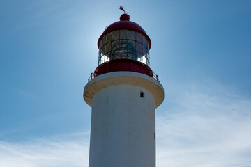A vintage lighthouse tower with a round red metal roof.  In the center of the lighthouse is a vintage lamp made of multiple pieces of glass. On top of the white tower is a red metal wind arrow.