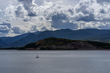 Fototapeta na wymiar Hillside and mountains with some fall foliage behind sailboat on Lake Dillon, Colorado, on partly cloudy late summer day with some sun shining through the clouds