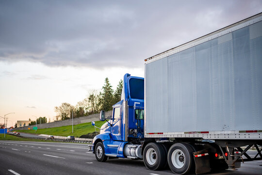 Bright blue day cab big rig semi truck with roof spoiler transporting cargo in dry van semi trailer running on the evening road