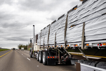 Powerful dark big rig classic semi truck transporting covered lumber wood on flat bed semi trailer...