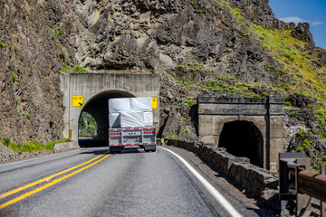 Professional industrial big rig semi truck with loaded flat bed semi trailer running on the winding road through a tunnel in the rock at Columbia Gorge area