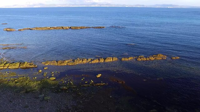 Aerial view of the sea side. Some rocks perfectly aligned on a narrow row are seen. Small ondulations seen on the water quite close to each other. The view moves backwards like a zoom out.