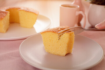 A piece of corn cake being cut by a fork, by a female hand, on a plate, on a table, with a cake and a cup in the background