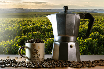 Coffee beans in a cup and Italian coffee maker on a wooden table with coffee field background in Brazil