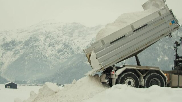 A large industrial truck dumping a load of snow onto a snow covered surface with a large mountainous landscape in the background - daytime.
