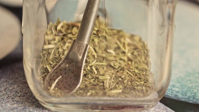 Pour in yerba mate green tea into a glass with bombilla inside. Healthy natural green drink preparation. Slowly adding drought. Putting yerba mate tea powder in a jar.