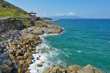 Along the slope of the mountain, along the seashore, a wooden footpath with rope railings winds. A canopy is visible in the distance. Turquoise waves foam on the stones. Clear blue sky. Vietnam