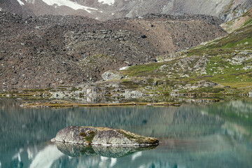 Rock in turquoise mountain lake. Snowy mountain reflected in azure clear water of glacial lake. Beautiful sunny background with snow-white glacier reflection in green water surface of mountain lake.