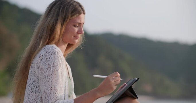 Young and beautiful caucasian woman sitting on the beach and using digital tablet for drawing the ocean picture for relaxing in the evening while travel at tropical country on summer holiday vacation.