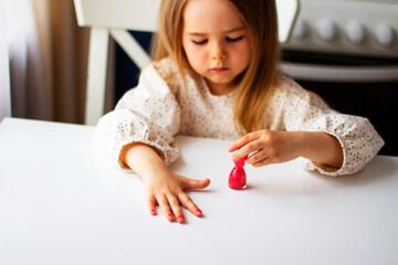 Little girl paints her nails with pink nail polish. Cute toddler girl playing with colorful nail polish doing manicure. Cosmetics and beauty concept.
