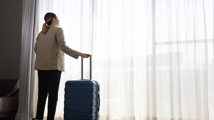 Young Asian girl in smart casual dress takes a breath moment at an apartment room window to inspire relax and stretch. Female visitor or traveler stands in front of white curtain look out for future.