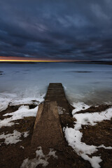 harsh winter evening landscape. old wooden pier on a deserted lake shore under a dramatic sky with a streak of sunset glow