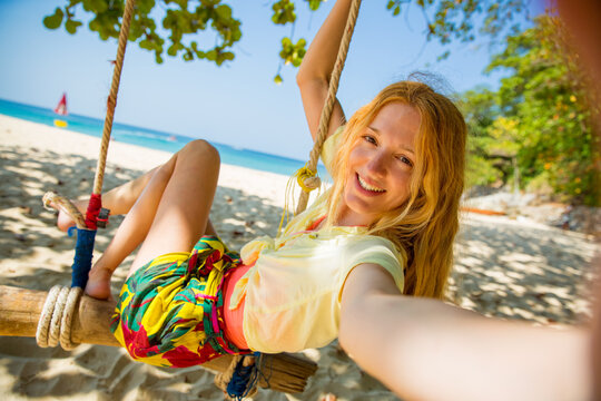 Happy young woman with red hair laughing and swinging on swing on a tree at the beach, taking a selfie. Beautiful summer sunny day, turquoise sea, white sand, tropic landscape. Phuket, Thailand.