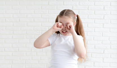 A girl in a white T-shirt covers her eyes with her hands and cries. White brick background. Copy space.