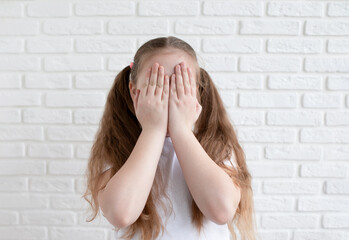 A girl in a white T-shirt covers her face with both hands. Game concept and puzzles. White brick background. Copy space.