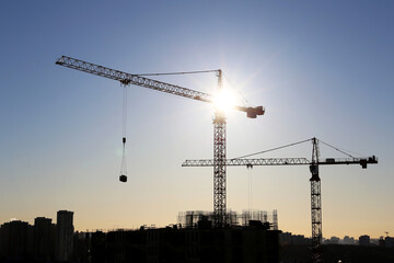 Silhouettes of tower cranes and unfinished residential building against the sky and shining sun. Housing construction, apartment block in city