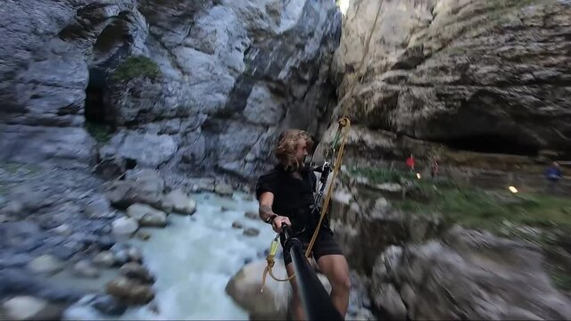 Young, Fit And Strong Man With Long Hair And Beard Jumps With A Smile Out In The Canyon With A Bungee Jump Rope Holding A Selfie Stick. Swings Back And Forth On An Adrenaline Rush Experience.