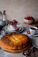 The round pie is cut into pieces. Inside, the filling is made of cherries and sugar. On a wooden table there are dishes, a cup of tea. Berries and flowers are laid out.