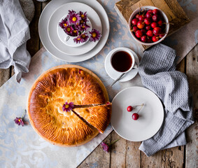The round pie is cut into pieces. Inside, the filling is made of cherries and sugar. On a wooden table there are dishes, a cup of tea. Berries and flowers are laid out.